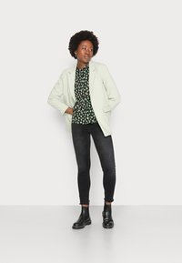 Blazer verde claro sobre uma blusa preta com padrão e detalhes verdes, combinado com jeans skinny pretos e botins pretos. Texturas suaves.