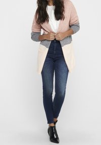 Femme portant un cardigan coloré en rose, gris et crème sur une blouse blanche, associé à un jean skinny bleu foncé et des bottines noires.