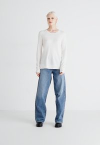 Pull blanc à manches longues et jean bleu ample. La tenue présente un design simple avec une texture douce et des accents minimaux, portée avec des bottes noires.