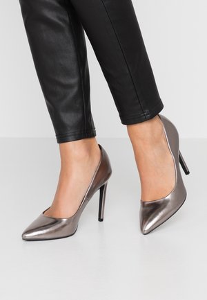 Des chaussures à talons hauts en argent métallique avec un bout pointu et une texture lisse et élégante. Portées avec un pantalon noir ajusté, laissant apparaître la cheville.