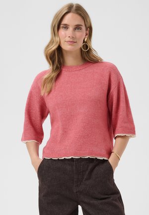 Femme aux longs cheveux ondulés porte un pull rose à manches courtes avec bordure en dentelle, pantalon foncé, boucles d’oreilles créoles dorées et bracelet, les mains dans les poches.