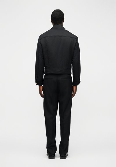 Άνδρας που στέκεται γυρισμένος προς τα πίσω, φορώντας μαύρο κοντό μπουφάν, μαύρο παντελόνι και μαύρα παπούτσια σε λευκό φόντο.