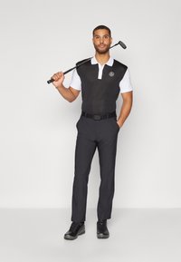 Čierny bezrukávový golfový pól s bielym golierom, skombinovaný s tmavými nohavicami a čiernymi topánkami. Model drží golfovú palicu vo vzpriamenej polohe.