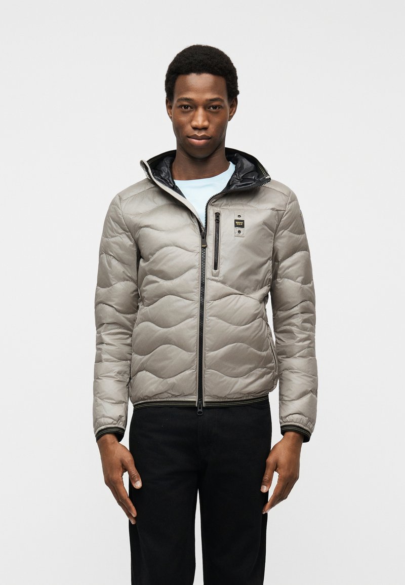 Άνδρας που φοράει ανοιχτό γκρι μπουφάν με φερμουάρ στις τσέπες, μαύρο παντελόνι και ανοιχτό μπλε πουκάμισο, μπροστά σε απλό λευκό φόντο.