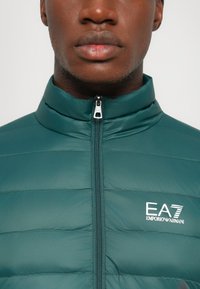 Nærbilde av en person som har på seg en teal polstret jakke med høy krage og glidelås, samt hvit EA7 Emporio Armani-logo på brystet.