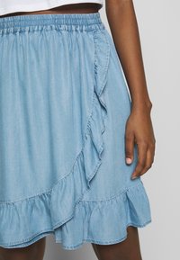 Jeansrock in Hellblau mit einem Rüschensaum und seitlicher Klappe. Verfügt über einen elastischen Bund für zusätzlichen Komfort. Ganze glatte Textur.