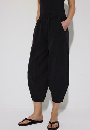 Osoba nosi voľné čierne krátke nohavice s elastickým pásom, ruky vreckách a čierne žabky, stojaca pri jednofarebnej stene.