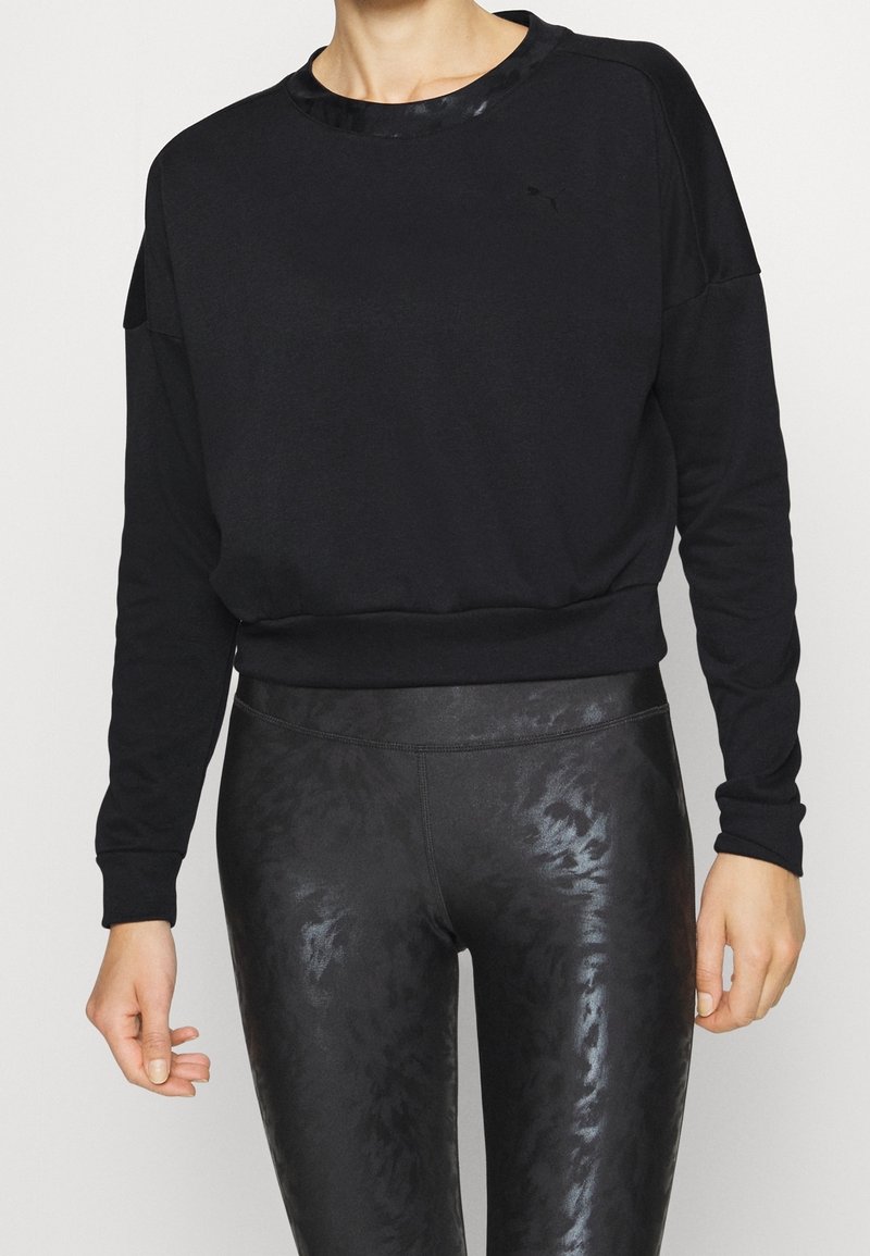 Femme portant un sweat-shirt noir à manches longues et des leggings noirs brillants à motifs, debout devant un fond clair.
