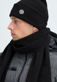 Nuori mies, jolla on päässään musta beanie, jossa on valkoinen logo, musta huivi, hopeiset renkaat ja harmaa vuorattu takki, jossa on vetoketjullinen tasku.