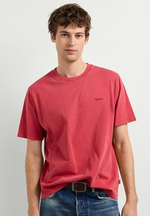 Joven con el pelo corto y castaño, vistiendo una camiseta roja de cuello redondo y vaqueros azules con un cinturón negro, sobre un fondo blanco liso.