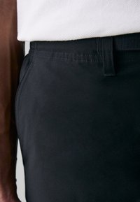Pantalon noir en tissu lisse, avec un devant plat, une taille standard et une couture visible le long des coutures.