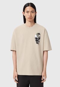 Camiseta de algodón beige con mangas cortas, que presenta un gráfico negro de una calavera con una serpiente, y un diseño de ajuste relajado.