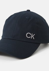Sötétkék pamut baseball sapka ívelt vízszintes éllel és ezüst színű hímzett "CK" logóval. A sapka élén varrott részletek és szellőzők találhatók.