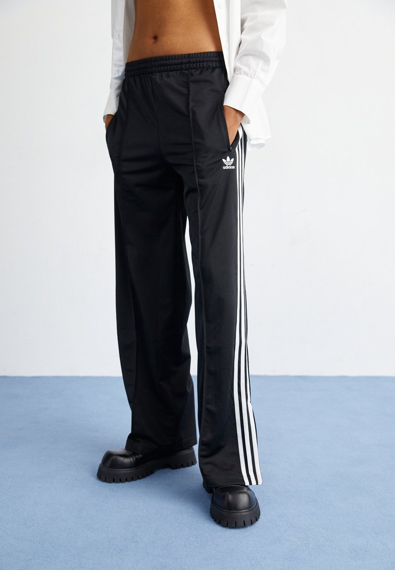 Black athletic pants with white side stripes, made of smooth fabric, featuring an elastic waistband and side pockets. Worn with chunky black shoes.