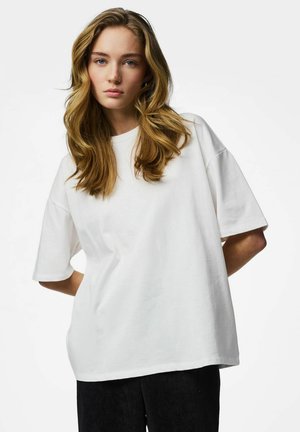 Noor naine pikkade laineliste blondide juustega, kandes üle suuruses valget T-särki ja musti pükse, valgel taustal.