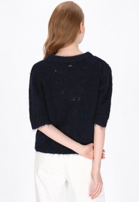 Granatowy sweter dzianinowy z krótkimi rękawami, luźny krój i teksturowany wzór. Posiada surowe wykończenie dołu i małe logo na zapięciu z tyłu.