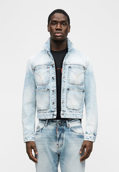 Άνδρας που φορά ανοιχτόχρωμο τζιν μπουφάν με δύο τσέπες στο στήθος και τζιν παντελόνι που ταιριάζει, στέκεται μπροστά σε απλό λευκό φόντο.