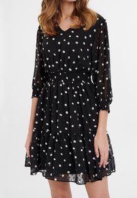 Robe noire à pois avec un col en V, des manches trois-quarts et une taille élastique. Jupe fluide à volants avec une texture translucide.