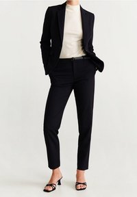 Czarna dopasowana marynarka nałożona na kremowy top, zestawiona z czarnymi wąskimi spodniami i strappy czarnymi szpilkami. Gładki materiał o eleganckim kroju.