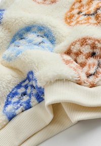 Nahaufnahme eines creamfarbenen Fleece-Stoffs mit erhöhten blauen und braunen kreisförmigen Mustern und geripptem Saum.