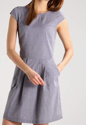 Kobieta ubrana w jasnoszarą sukienkę o krótkich rękawach sięgającą do kolan, z dopasowaną talią i bocznymi kieszeniami, stojąca na tle gładkiego, jasnego tła.