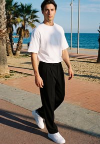 Mann trägt weißes T-Shirt, schwarze Hose und weiße Turnschuhe, geht auf einer Strandpromenade mit Palmen und Meer im Hintergrund.