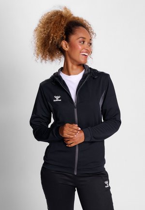 Black athletic jacket with zip closure, hood, and gray accents. Soft fabric with a textured pattern. Logo on left chest. Paired with black pants.