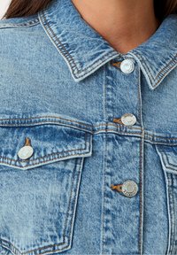 Světle modrá džínová bunda s klasickým límcem, stříbrnými knoflíky, kontrastním šitím a náprsními kapsami. Disponuje vybledlým, strukturovaným povrchem.