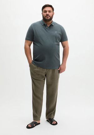 Mężczyzna stoi z rękami w kieszeniach, ubrany w ciemnozieloną koszulkę polo, oliwkowe spodnie i czarne sandały na jednolitym białym tle.