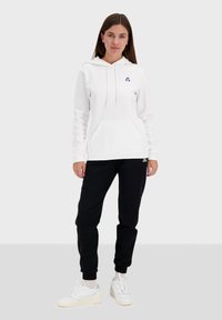 Felpa bianca con tasca anteriore e cordino, caratterizzata da un piccolo logo ricamato. Abbinata a pantaloni sportivi neri e scarpe da ginnastica bianche.