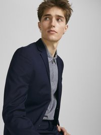 Marineblauer Anzugjacke über einem hellgrauen Strickshirt. Die Jacke hat eine taillierte Passform und einen subtilen Glanz. Das Model hat kurze Haare und silberne Creolen.