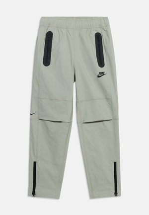 Pantalon Nike gris clair avec taille élastique, poches zippées noires sur les cuisses, rabats aux genoux et zips aux chevilles. Logos Nike noirs sur la cuisse et le mollet.