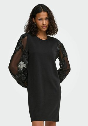 Femme portant une robe noire longueur genou avec des manches bouffantes transparentes brodées de fleurs, debout devant un fond clair uni.