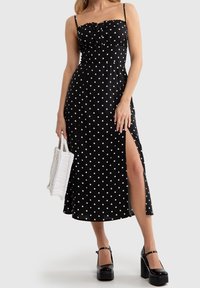 Kvinna i en svart midiklänning med vita prickar och hög slits, som håller i en vit vävd väska och har på sig svarta platåskor.