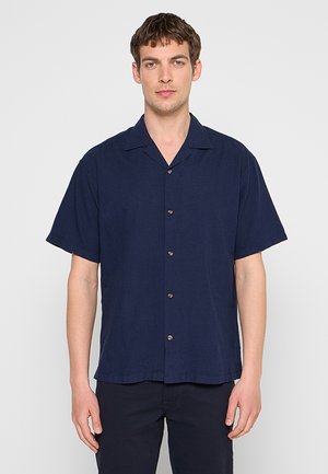 Jeune homme portant une chemise bleu foncé à manches courtes, debout devant un fond gris clair uni.
