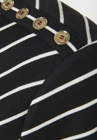 Mustavalkoinen raidallinen kangas, jossa on kultaiset napit ja punaisia yksityiskohtia, esittelee pehmeää tekstuuria ja klassista muotoilua.