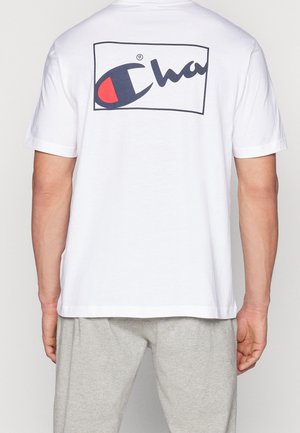 Mężczyzna w białej koszulce z krótkim rękawem z dużym logo Champion w kolorze granatowym i czerwonym na górnej części pleców, w połączeniu z jasnoszarymi spodniami.