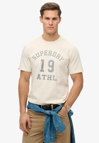 Camiseta de algodón beige con gráficos grises "SUPERDAY 19 ATHL." Modelo lleva pantalones caqui, con un denim azul atado a la cintura y un reloj visible en la muñeca.