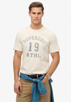Beige bomulds T-shirt med grå grafik "SUPERDAY 19 ATHL." Model iført khaki bukser, blå denim bundet om taljen, håndledsur synlig.