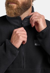 Pullover in pile nero con colletto alto, zip a metà e polsini elasticizzati. Presenta un piccolo logo sul petto e una superficie in tessuto testurizzato.