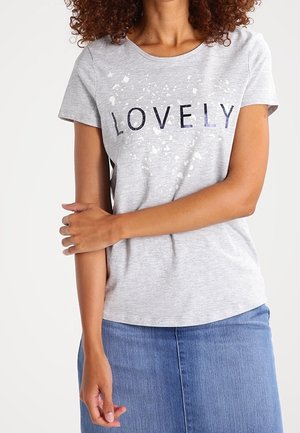 Nainen kiharat hiukset, pukeutunut vaaleanharmaaseen T-paitaan, jossa on "LOVELY"-teksti ja valkoinen roiskekuvio, yhdistettynä siniseen farkkuhameeseen.