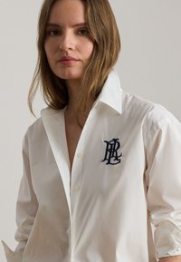 Młoda kobieta z długimi brązowymi włosami, ubrana w białą koszulę zapinaną na guziki, z haftowanym granatowym monogramem na lewej piersi.