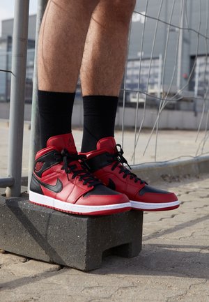 Person iført røde og sorte høje sneakers med sorte sokker står på en betonblok udendørs nær et metalgelænder.