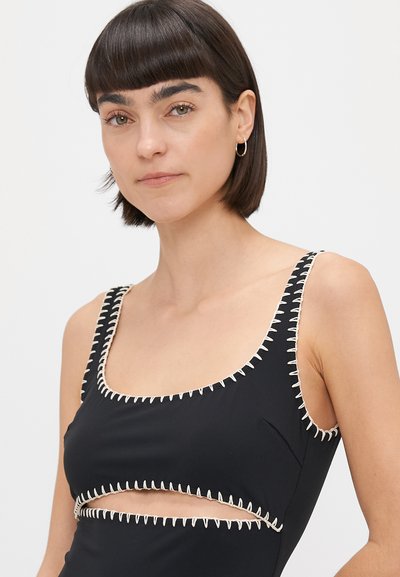 Čierne plavky s hlbokým výstrihom a výrezom. Obsahujú kontrastné béžové stehy pozdĺž okrajov, čím vytvárajú textúrovaný vzor.