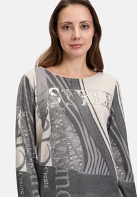 Graues und beiges Langarmtop mit einer Vielzahl von grafischen Drucken und Verzierungen, darunter Text und Muster, mit einem weiten Ausschnitt.