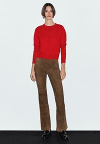 Červený pletený cardigan s detaily knoflíků, spárovaný s hnědými semišovými kalhotami do zvonu. Modelka v černých mokasínech, stojící na jednobarevném pozadí.