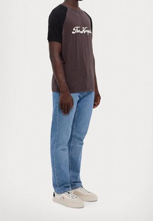 Man draagt een zwart-bruin raglan-T-shirt met witte tekst, lichtblauwe spijkerbroek en witte sneakers, staand zijwaarts tegen een effen achtergrond.