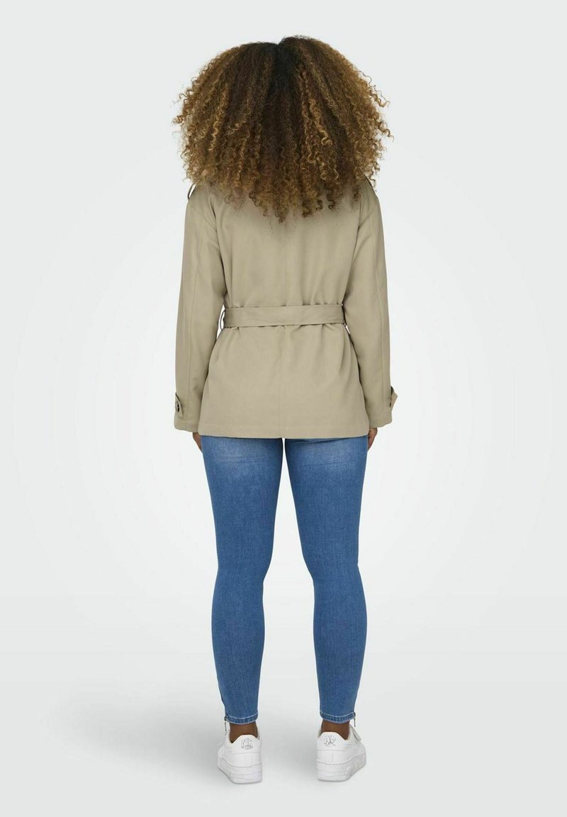Veste beige à ceinture avec manches longues, présentant une coupe droite, portée sur un jean bleu et des baskets blanches. Tissu texturé avec poches latérales.