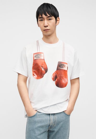 Λευκό μπλουζάκι με τύπωμα κόκκινων γαντιών πυγμαχίας με ετικέτες "MOSCHINO", σε συνδυασμό με τζιν παντελόνι ανοιχτού γαλάζιου χρώματος.