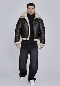 Giacca di pelle marrone con colletto e polsini in shearling, abbinata a un dolcevita nero a coste e jeans scuri dalla vestibilità ampia. Sneakers bianche.